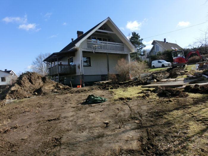 Ett hus med upplyft veranda och en grävmaskin arbetsområde med uppkörd jord, indikation på dräneringsarbete.