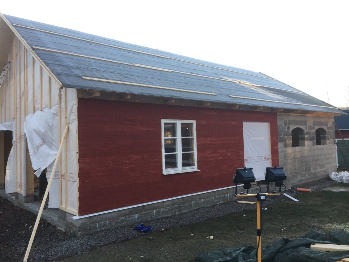 Nypanelad röd byggnad med plåttak och treglasfönster, läkt på taket, och garageport under konstruktion.