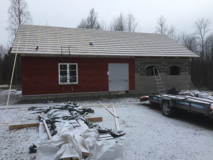 Byggprojekt av en röd träbyggnad med takläkt och en påbörjad takkonstruktion, omgiven av snö och byggmaterial.