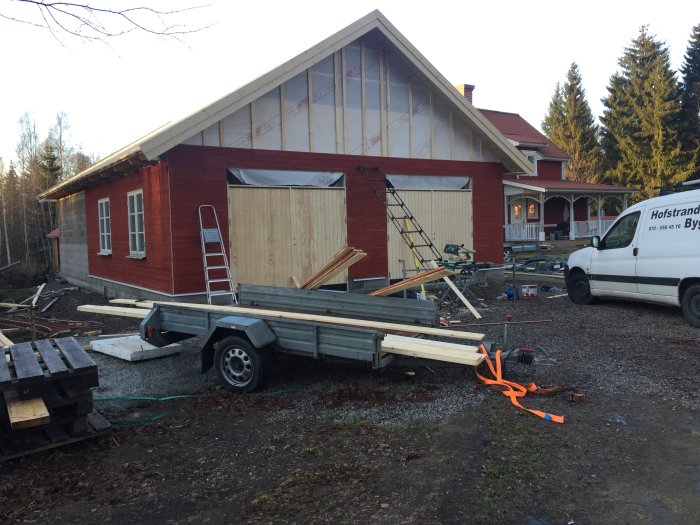 Delvis färdigställd röd byggnad med träpanel, byggmaterial och verktyg spridda framför, samt vita fönsterkarmar.