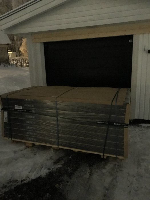 Pallar med förpackad ekparkett står utomhus i snö framför en garageport i dunkel belysning.