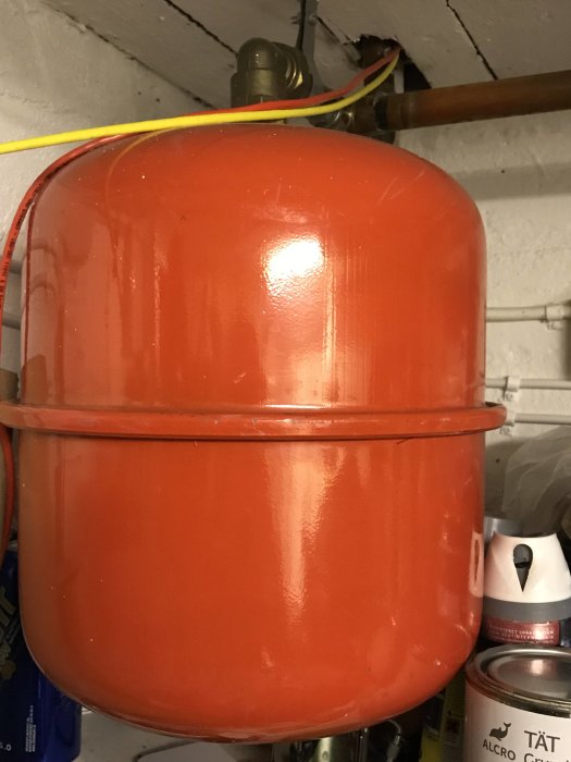 Ett rött expansionskärl i en källare med rörkopplingar och ledningar i bakgrunden.