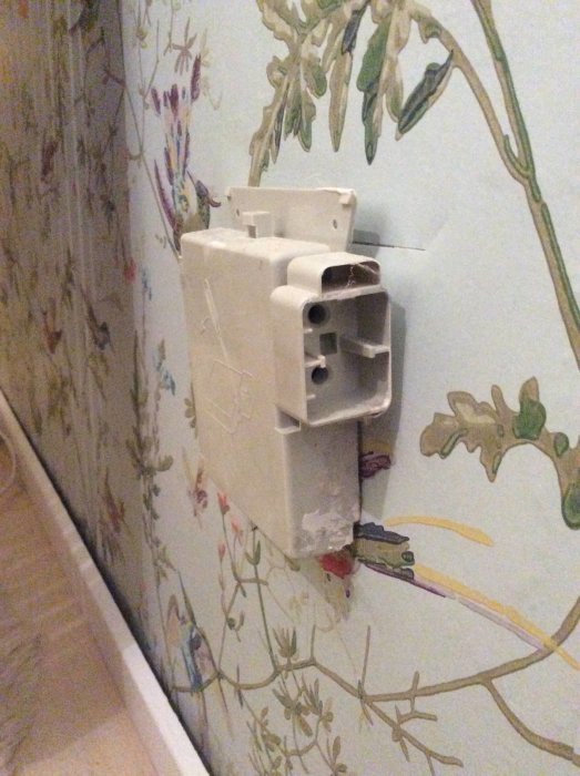 Äldre väggmonterad radiatoranslutning utan radiator, synliga ledningar och tapeter med blommönster i bakgrunden.