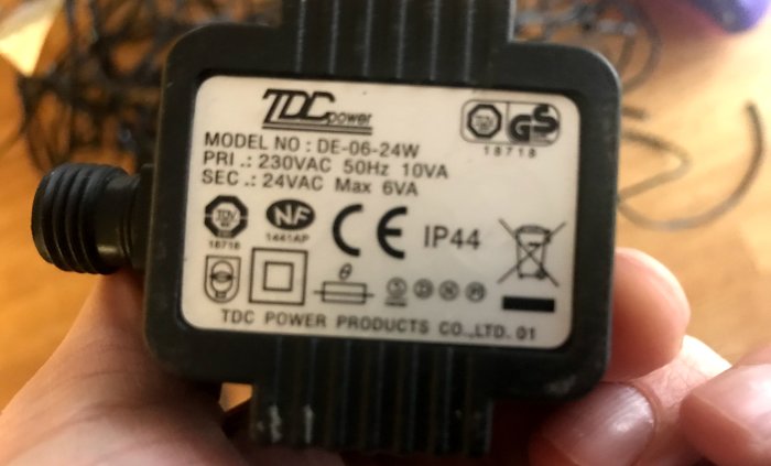 Hand håller en transformator med etikett som visar modellnummer, elektriska specifikationer och säkerhetsmärkningar.