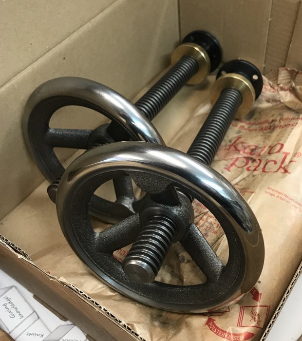 Gjutjärnsrattar och trapetsgängad stång ligger på kartongfyllnad i en kartong.
