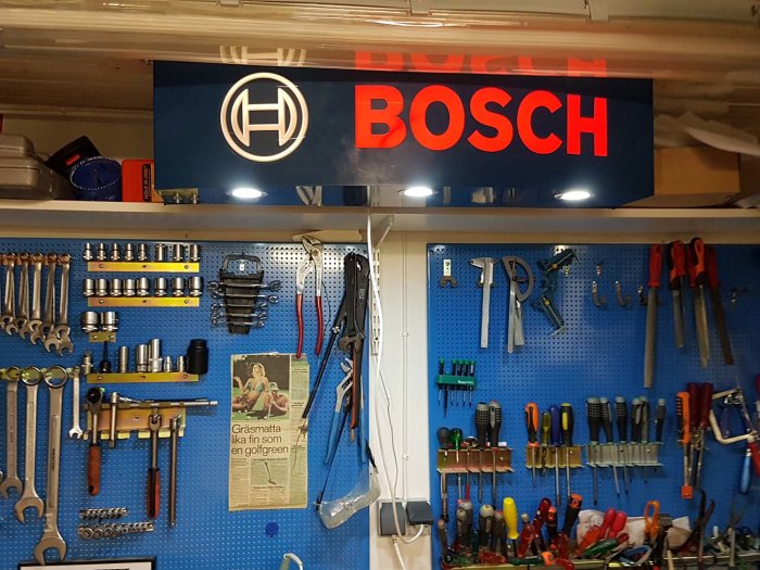 Vägg i garage med upplyst skylt märkt 'BOSCH' ovanför pegboard organiserad med olika verktyg och arbetsbordsbelysning.