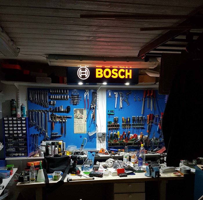 Garage med upplyst Bosch-skylt ovanför arbetsbord med verktyg och rörigt bord.