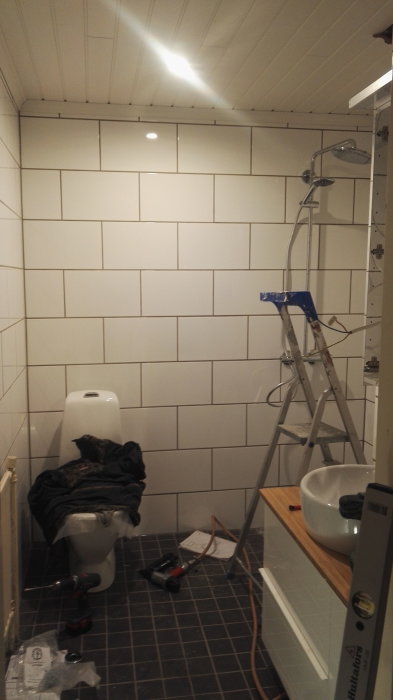 Badrum under renovering med vita kakelväggar, toalett täckt med byggmaterial, och en stege.