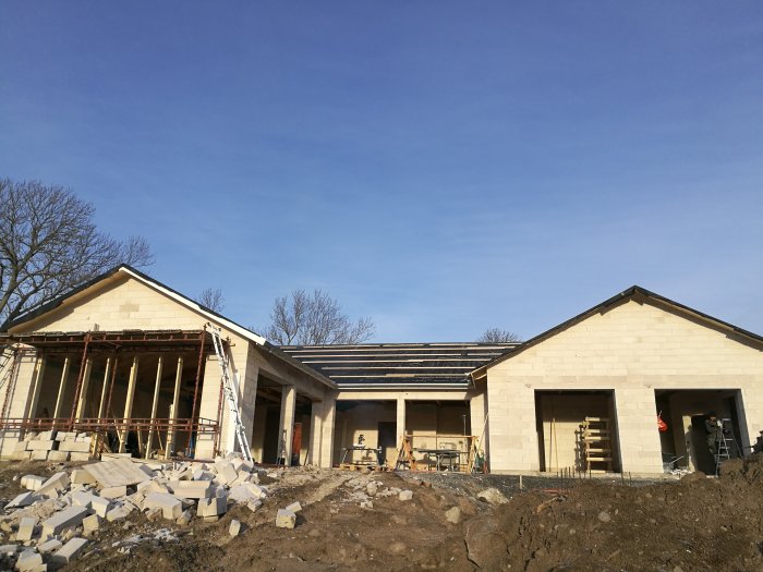 Byggarbetsplats med pågående arbete på ett hus under en klarblå himmel, med synliga byggmaterial och stegar.