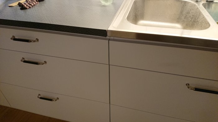 Köksinredning med vita lådfronter och handtag, svart bänkskiva och rostfri diskho.