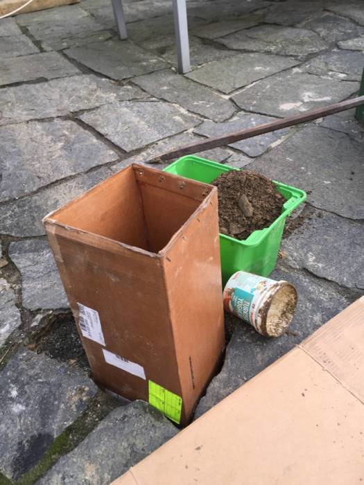 En kartong används som gjutform för plint med grön hink fylld av grus och en övergiven spade i bakgrunden.