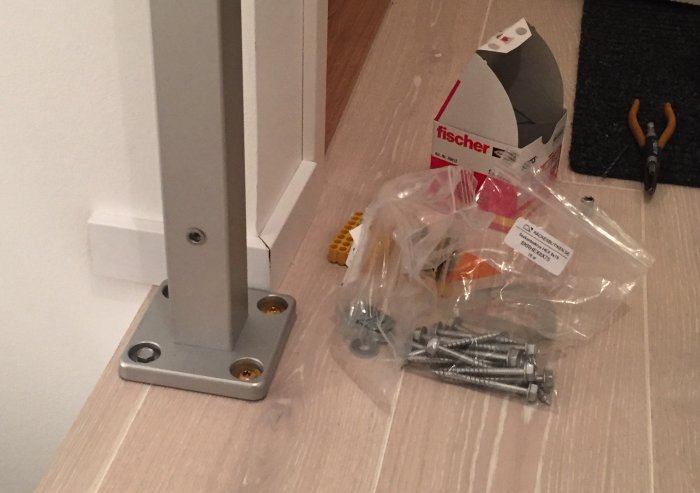 Räjähtänyt kuva asennusprojektista, jossa on metallinen seinäkiinnike, ruuveja ja työkaluja lattialla.