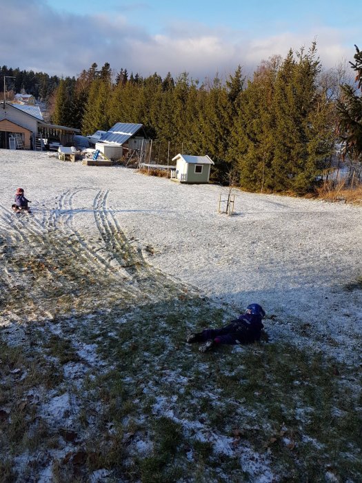 Två barn leker i en tunn hinna av snö på en sluttning, med träd och en lekstuga i bakgrunden.