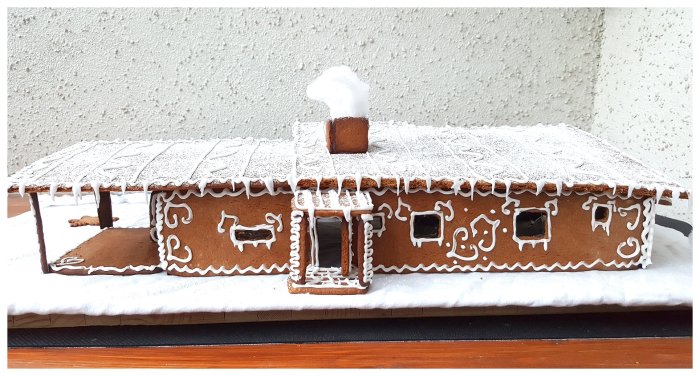 Handgjort pepparkakshus med detaljerat dekor och snötäckt tak mot en betongvägg.