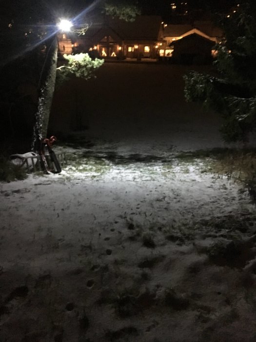 Snöig backe upplyst av en LED-strålkastare med trädgren i förgrunden och hus i bakgrunden nattetid.