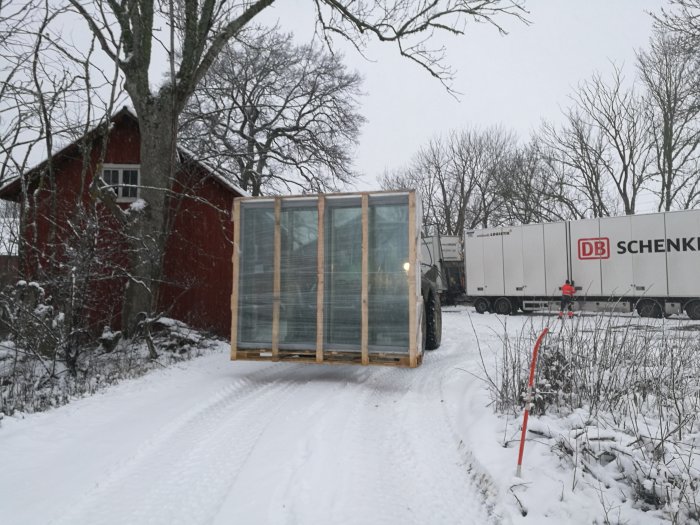 Nya fönster inpackade på en pall framför en lastbil i ett snöigt landskap med ett rött hus i bakgrunden.