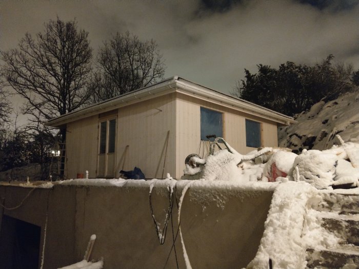 Ett hus under vinter med snöbelagd byggarbetsplats i förgrunden nattetid.