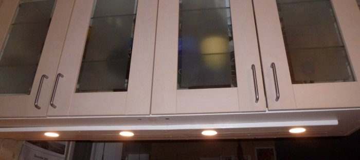 Ett vitlackat frihängande vitrinskåp med glasdörrar och synliga skruvskallar, upplyst av fyra underspotlights.