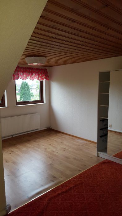 Ett tomt rum i ett äldre hus med trägolv, vitmålade väggar och träpanel i taket, med ett fönster som släpper in dagsljus.