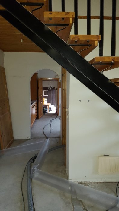Bild inifrån ett hus under renovering, visar en trätrappa och blickar genom en bågformad dörröppning mot ett annat rum.