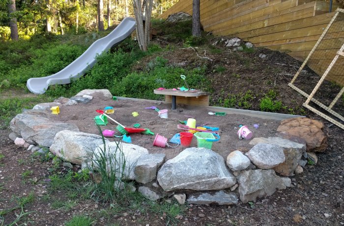 Nybyggd lekplats med sandlåda, leksaker och rutschkana omgiven av stenar och natur.