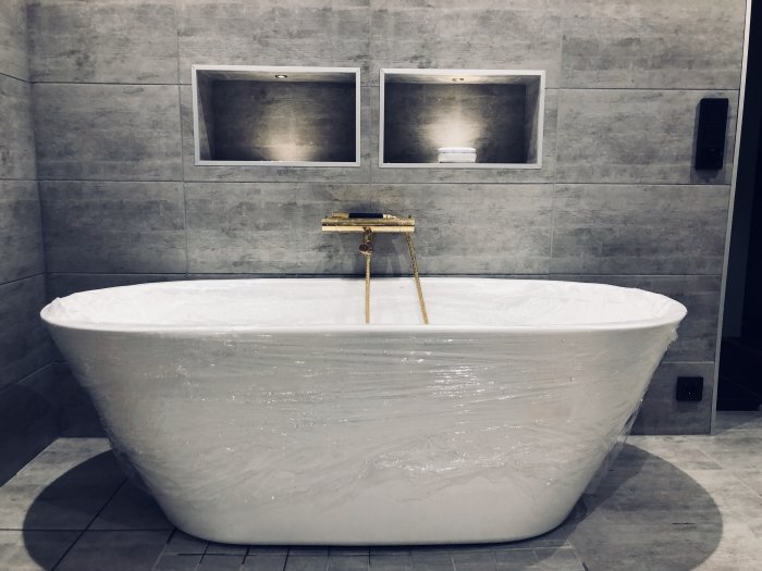 Nytt fristående badkar i plastfilm med monterad guldkran i modernt badrum.