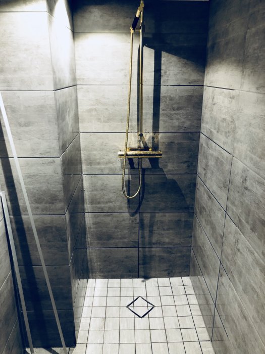 Guldfärgad duschblandare monterad i en grå kaklad duschhörna med öppen glasdörr.