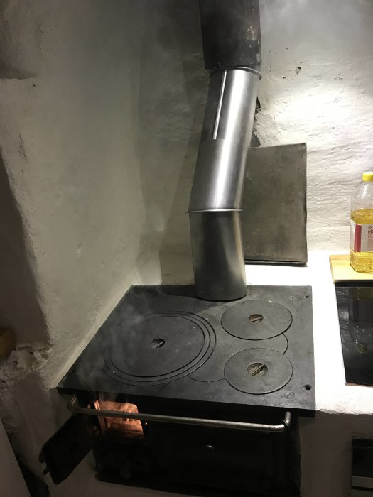 Järnspis i ett kök med påslagen eld och anslutna rökrör upp mot den vitmålade väggen, utan synligt drag.