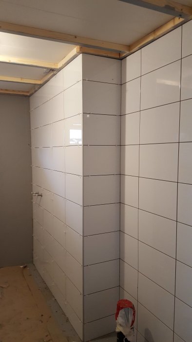 Renoveringsfas med kaklade vita väggar i ett badrum under uppbyggnad, skydd och verktyg synliga.