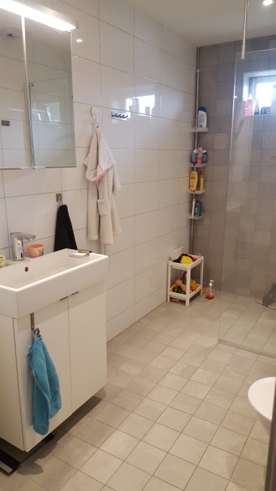 Modernt badrum med vita kakelväggar, grått golv, och duschhörna med glasdörr.