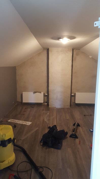 Renovering av ovanvåning med skråtak, synlig betongvägg, radiatorer och verktyg på trägolv.