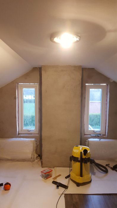 Renovering av ovanvåning med två fönster, spacklade väggar, konstruktionsmaterial och en golvdammsugare.