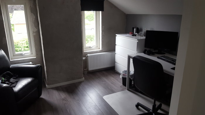 Renoverad ovanvåning med gråa väggar, skrivbord med dator och svart fåtölj.