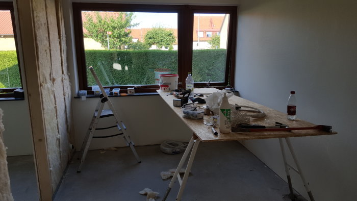 Rum under renovering med verktyg och material på ett arbetsbord framför ett fönster.