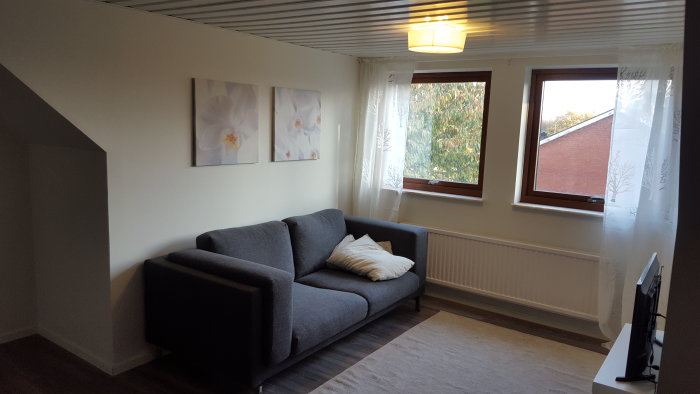 Renoverad ovanvåning med grå soffa, vita orkidétavlor, trägolv och två fönster med utsikt.