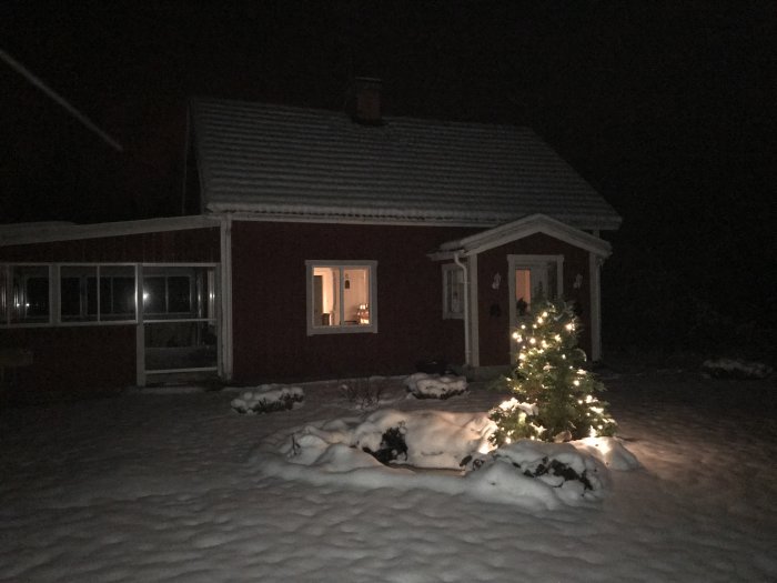 Röd stuga med snötäckt tak och ljusslinga på ett julpyntat träd, omgiven av snö i skymningen.