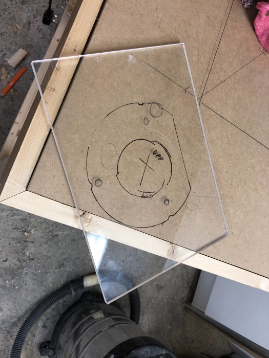 Plexiglasskiva med uppritat mönster av fräsplattan ligger på ett träbord, redo för bearbetning.