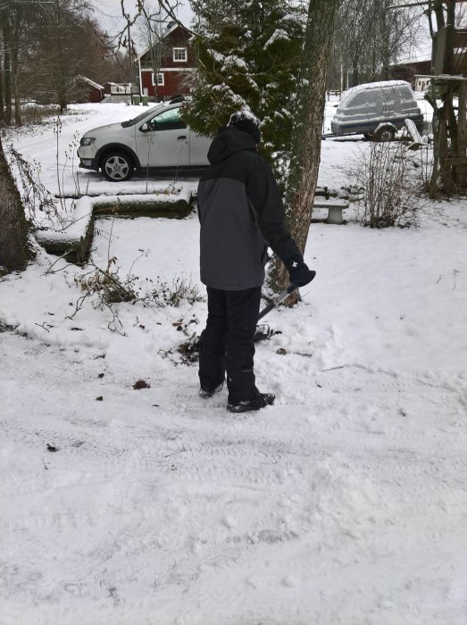 Person använder manuell snöslunga på uppfart med snötäckt landskap i bakgrunden.