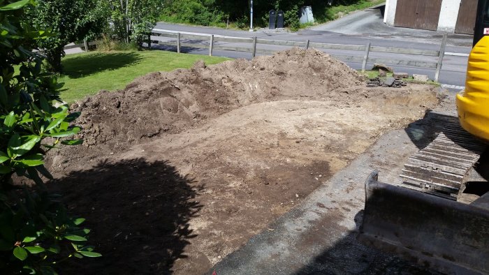 Uppgrävd trädgårdsyta med en hög jord i förgrunden och en grävmaskin till höger, i ett pågående anläggningsprojekt.