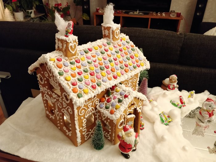 Detaljerat pepparkakshus med färgglada dekorationer och kristyr, omgivet av julfigurer och "snö