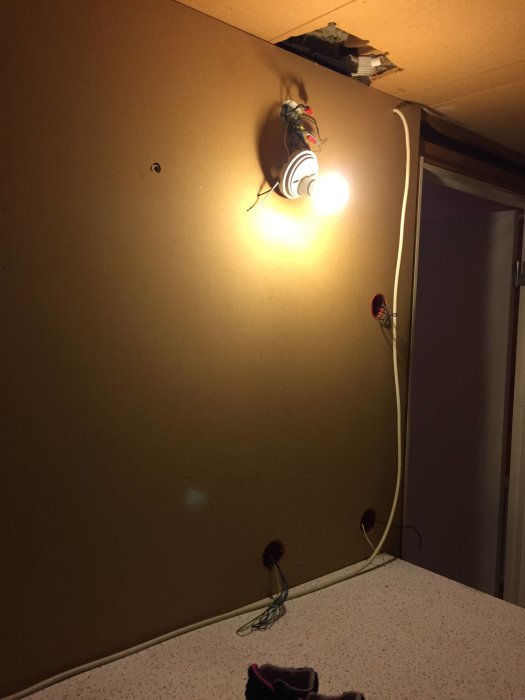 Ofärdigt rum med spånskivevägg och exponerade elinstallationer, där en glödlampa tänds och kablar hänger löst.