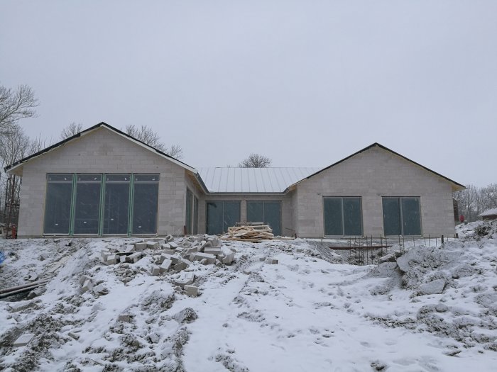 Nybyggt hus med tak och fönster installerade, täckt av snö, byggmaterial framför.