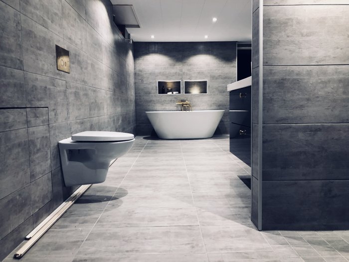 Modernt badrum med grå kakelväggar, fristående badkar och toalett, vy från tvättstuga.