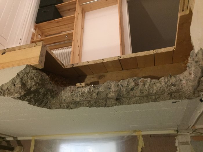 Brudt betonvæg og træstruktur i et hjem under renovering, med udsigt til spån og gulvbjælker ovenover.