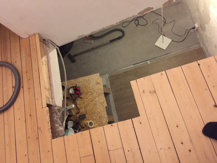 Et udsavet hul i et trægulv over et rum med værktøj og materialer. En støvsugerslange og en lampe ses også, der ligner et renoveringsprojekt.