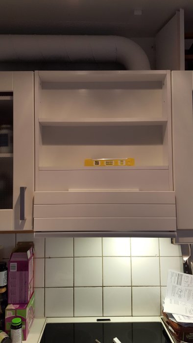 Mörkt köksutrymme med en vit ventilationskanal ovanför skåpet och en vattenpass på skåpets hylla.