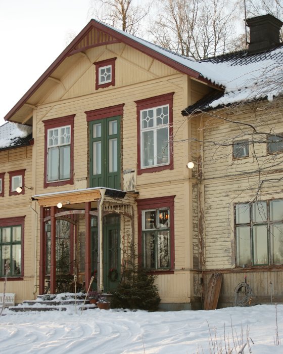 Snötäckt traditionellt svenskt trähus med tända kakelugnar synliga genom fönstren och juldekorationer utanför.