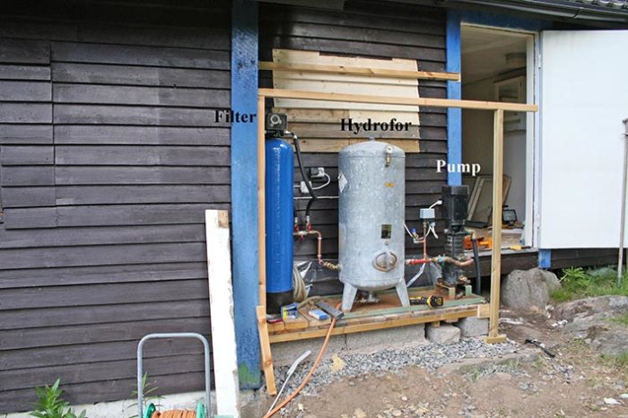 Installation av vattenpump, hydrofor och filter vid en husvägg, under pågående arbete.