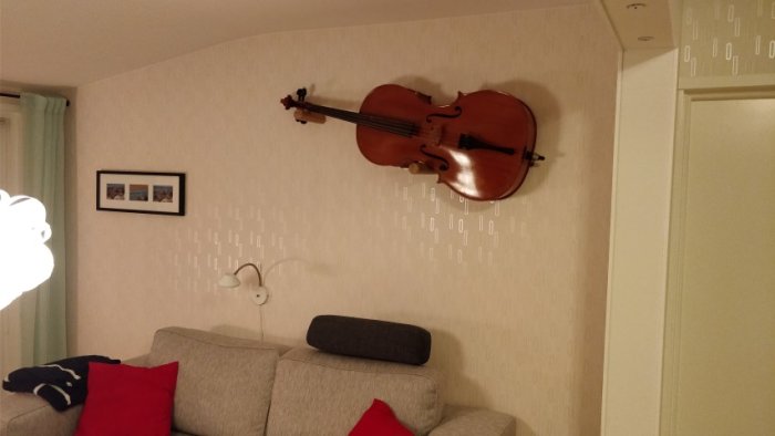 Cello upphängd på en tapetserad vägg över en soffa, med tillbehör för stabilitet.