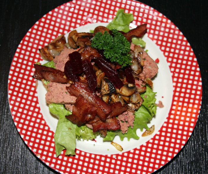 Ett smörrebröd med leverpastej, bacon, rödbetor och stekt svamp på ett rutmönstrat fat.